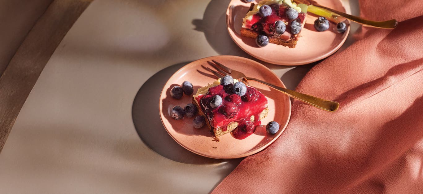 Two pale pink plates, each with a Blueberry Cheesecake Bars and garnish of fresh blueberries